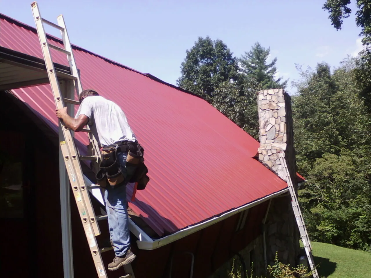 Roofer on a ladder beside a red metal roof and stone chimney — Soffit & Fascia Repair by Roofing Brothers in Lowman